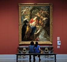 Two young women sit in front of a painting by Rubens of a group of women surrounded by putti