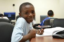 A child writing in a composition book and smiling at the camera