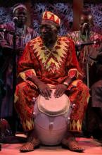 A man in a brightly colored red and yellow outfit plays a large drum with his hands.