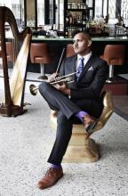 a youngish bald African American man sits on a chair with a trumpet in his lap and a harp in the background