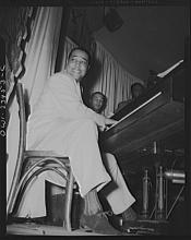 Duke Ellington seated at a piano smiling at someone offscreen as he plays