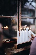 the hands of two people as they light a wood block menorah that's set on a window ledge