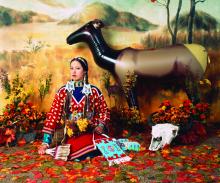 Woman in Native outfit sitting in front of a plains background  with a cow skull next to her and an inflatable elk behind her. 