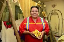 Standing in front of some of the tools of his craft, Gerald Hawpetoss smiles and holds a pair of small moccassins and an example of his beadwork.