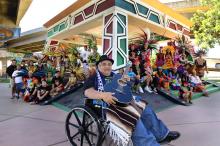 Ramón "Chunky" Sánchez sits in a wheelchair, holding a guitar and smiling, in front of a large group of communitiy members and performers wearing colorful and elaborate Aztec costumes.