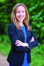 White woman wearing a blue dress and a dark blue blazer, standing in front of a wooded backround