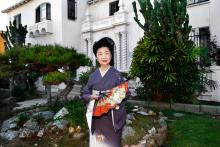 A Japanese woman holding a fan poses for a photo in front of a large white home