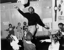 A Black man in a black shirt is dancing around in front of children in a classroom.