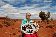 An elderly woman with glasses sits in the high desert holding two handwoven Navajo baskets. The red clay soil stands against the stark landscape and bright sky streaked with clouds. 