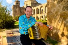 A man in a Southwestern blue printed shirt sits outside holding a gold-colored button accordion on his lap.  