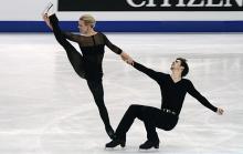 Female ice dancer holding her leg in the air while holding the hand of a mail ice dancer, who is in a sitting position. Both wear all black.