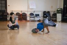Student wearing hat dancing while a girl sitting cross-legged nearby watches. 