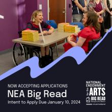 Two women in wheelchairs talk to one another. A table with a stack of books is between them.