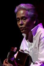 Man in white shirt with white hair brushed back playing guitar on stage. 