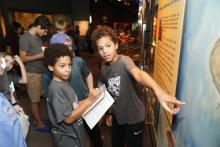 Two boys engage in an art exhibit at a museum. In the foreground, one child pointing at an information board while another take notes. 