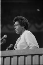 Barbara Jordan, an older Black woman wearing glasses, seen from the side as she speaks from behind a podium