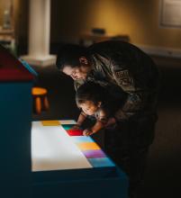 A father dressed in a military uniform holds his young son at an interactive art exhibit. The background is dark and there is light shining up illuminating their faces