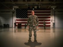 A man in a military uniform stands inside an aircraft hanger facing the camera with a giant American flag hanging in the background. 