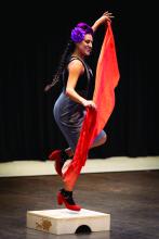 Woman with long braided hair and purple flower over her ear dancing on stage with a long orange scarf