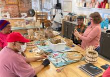 A small group of adults sit at a work table creating medium-sized baskets in an indoor studio setting with natural light.