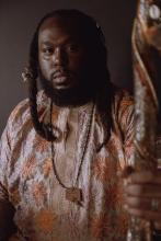 Portrait of Black man with beard and long braided hair wearing a orange flowered shirt and holding a staff.