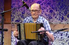 An elder man with glasses sits playing a button accordion with microphones around him and a colorful backdrop 