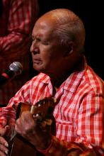 Balding Hawaiian man in red checkered shirt playing a uke. 