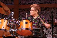 Older woman with short brown hair in colorful shirt playing drums. 