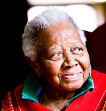 An older woman with short white hair and kind eyes looks off to the side and smiles, as if recalling a beautiful story. 