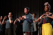A group of people standing looking off to the left while holding their hands at their chest, forming a shape of a heart.