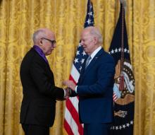 Older white male in blue suit posing with white man wearing glasses in black suit in front of flags and gold curtain.