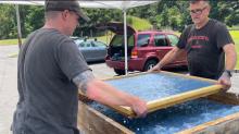 Two men stand outside under an awning holding a frame horizontally over a shallow box