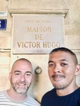 Man (left) wearing a striped shirt and man (right) wearing a dark blue shirt standing in front of a tan building that has the verbiage "VILLE DE PARIS MAISON DE VICTOR HUGO"