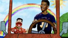 A smiling man sits next to a puppet while they drive in an animated car