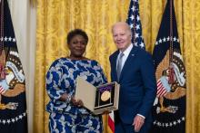 Older white male in blue suit posing with Black woman in blue floral dress in front of flags and gold curtain. 