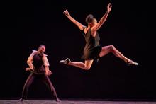 While a male-appearing dancer executes a move in the background, in the foreground a female-appearing dancer executes a leap.