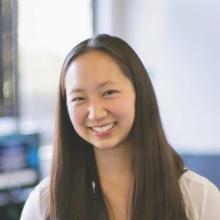 a headshot of Jenn Chang, who is an Asian-American woman with long dark hair parted in the center and falling over shoulders. She is smiling. The background behind her is blurred.