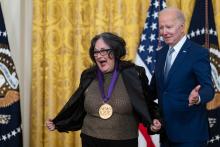 Older white male in blue suit posing with Hispanic woman in glasses wearing a black jacket in front of flags and gold curtain.