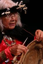 Native woman with white hair wearing headdress and holding up basket. 