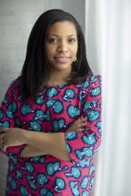 Pictured: Kellee Edusei, a butterscotch-skinned woman, is facing the camera, smiling, with her arms crossed. She has straight black hair and is wearing a pink and blue floral top.