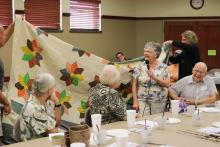 An older white woman stands at a table with a microphone in front of her handmade quilt