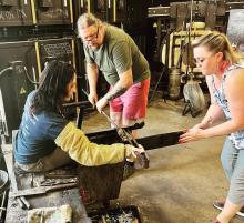 A group of three people uses metal tools to refine a piece of glass-blown art.