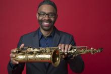 Black man wearing glasses and skullcap in blue shirt holding saxophone sideways in front of red background.