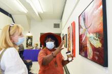 •	A woman wearing a facemask and hat discusses her artwork with a bystander at a gallery exhibit.