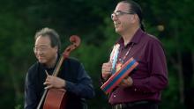 An Asian man in black shirt playing a cello next to a Native man in a purple shirt playing a drum.