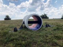 A barrel shaped sculpture with pink and purple interior sits in a grassy field