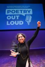 A young woman with two long braids holds a trophy in one hand with her other hand in the air. Behind her is a screen with the Poetry Out Loud logo.