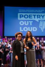 Two women, one middle aged and one teenaged, stand side by side smiling. The teenager holds an award