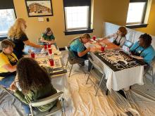 A group of 6 people sit around two tables painting book covers on large pieces of posterboard