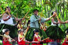 Dancers wearing grass skirts and floral wreaths holding bamboo sticks perform a traditional Hawaiian dance. 